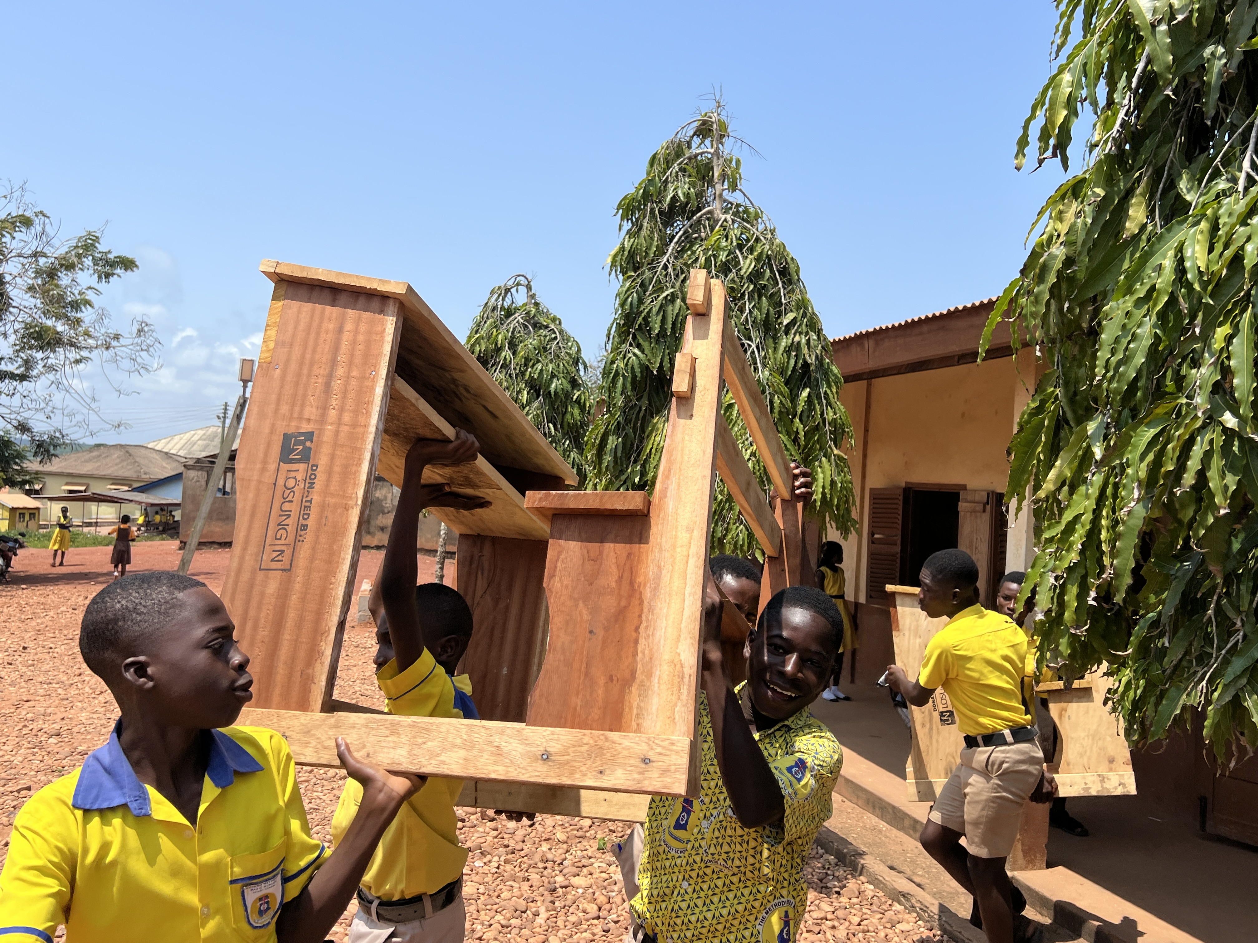 LÖSUNG N Klassenzimmerausstattung für Kinder in Ghana Kinder in Afrika tragen ihre gerade angelieferten Tisch-Stuhl-Kombinationen in den Klassenraum